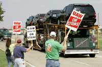 Un camión con coches nuevos sale de la planta de General Motors en Arlington, Texas, mientras empleados mantienen la huelga y se manifiestan con carteles en la entrada de la fábrica