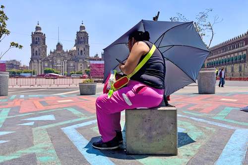 Una mujer toma un descanso en la Plaza de la Constitución para atender alguna llamada, mensaje o ver algún video en el teléfono celular, y pese a portar una sombrilla, ignora las recomendaciones oficiales de protegerse de los rayos solares.
