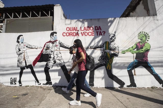 Jóvenes caminan en calles de Sao Paulo, Brasil, frente a un mural que representa la lucha de médicos contra el Covid-19 y Jair Bolsonaro. Foto Ap
