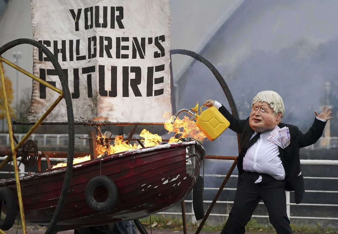 Un integrante de la campaña medioambiental Ocean Rebellion vestido como el primer ministro británico Boris Johnson prende fuego a la vela de un pequeño bote que dice "El futuro de sus hijos", durante una protesta cerca del sitio donde se llevará a cabo la COP26, el 27 de octubre de 2021. Foto Ap