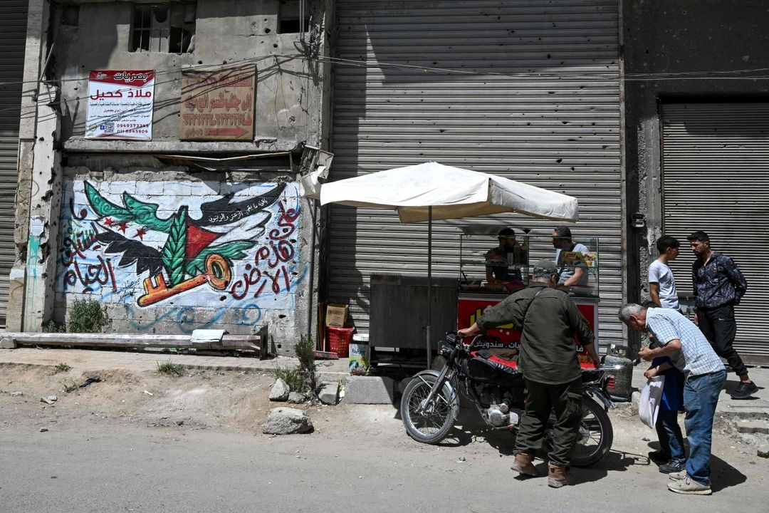 Hombres revisan una motocicleta junto a un mural con la imagen de las banderas siria y palestina, con una llave que simboliza un hogar palestino perdido en lo que hoy es Israel, en el campamento de refugiados palestinos de Yarmuk, al sur de Damasco, el 22 de mayo de 2025. Foto 