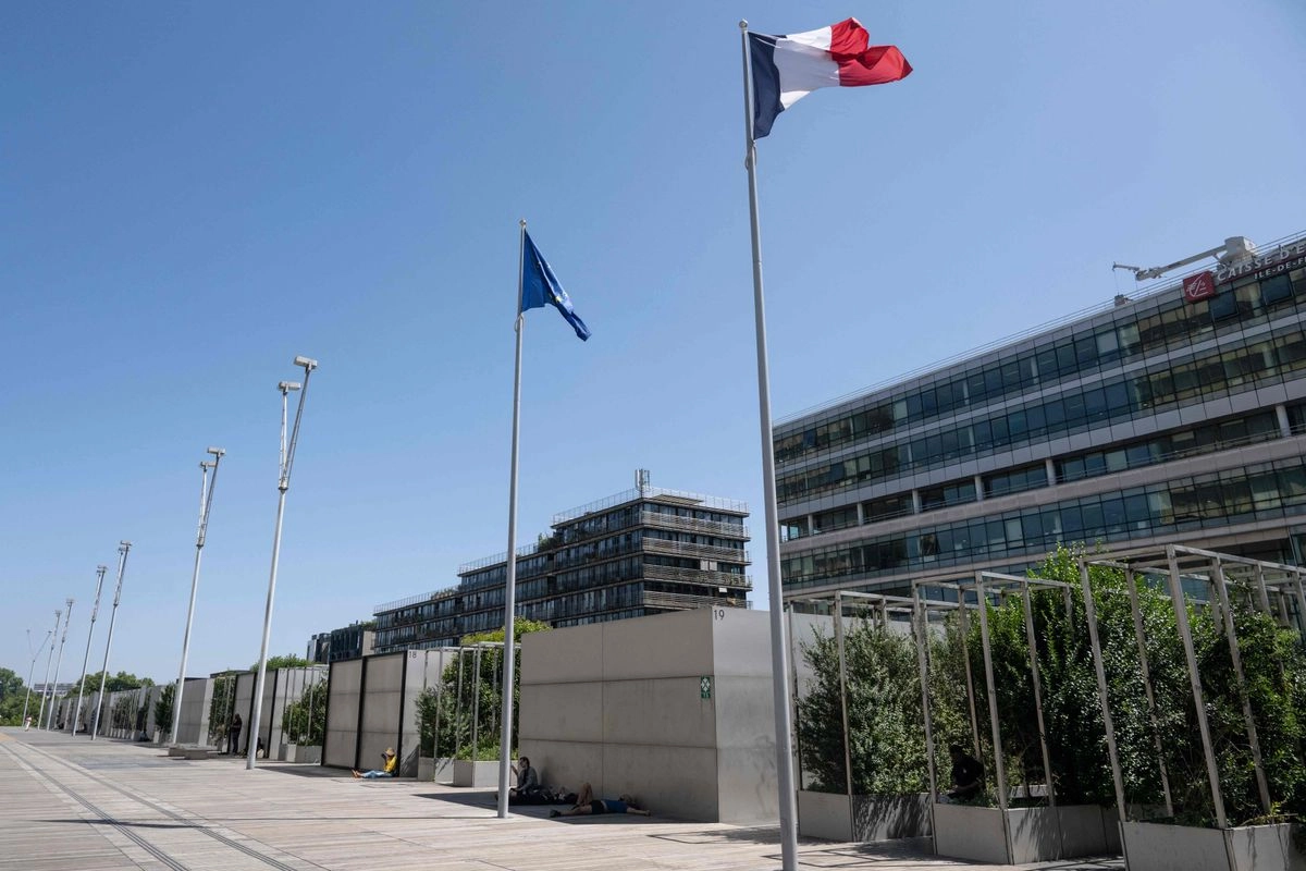 Personas sentadas a la sombra junto a una bandera francesa ondeando cerca de la Biblioteca Nacional de ese país, el 11 de agosto de 2025. 