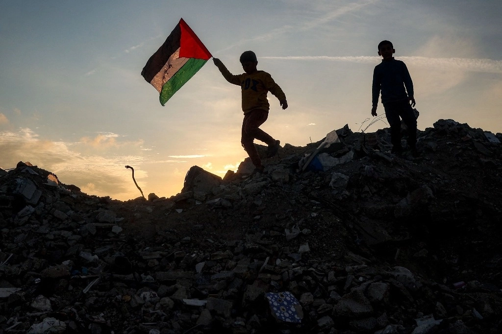 Un niño corre con una bandera palestina sobre un montón de escombros en un campamento para desplazados por el conflicto en Bureij, en el centro de la Franja de Gaza, el 17 de enero de 2025, tras el anuncio de una tregua en medio de la guerra en curso entre Israel y Hamas. Foto Afp