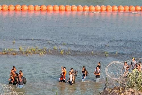 Migrantes caminan entre una valla de púas y una cadena de boyas en la línea divisoria con México en Eagle Pass, Texas. Ap