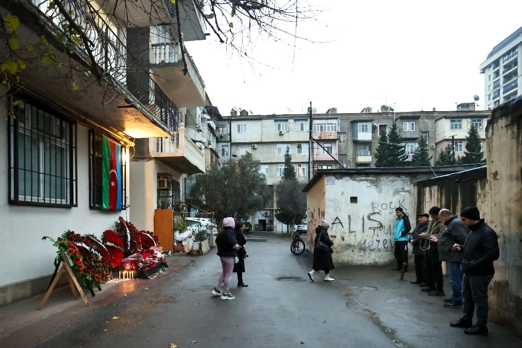 La gente se para frente a la pared de un edificio de apartamentos cubierto con una bandera nacional de Azerbaiyán, flores y velas en memoria del piloto Alexander Kalyanin, una de las víctimas que murieron en el accidente del Embraer 190 de Azerbaijan Airlines cerca del aeropuerto de Aktau, en Bakú, Azerbaiyán, el 28 de diciembre de 2024. Foto Ap