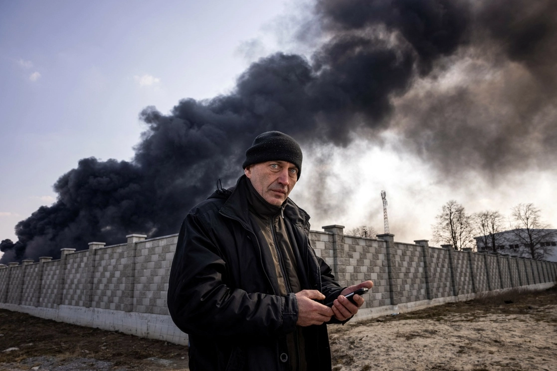 Un hombre sostiene un teléfono móvil mientras al fondo se levanta una columna de humo tras un ataque ruso a un depósito de combustible en la ciudad de Kalynivka, Ucrania, 182 kilómetros al suroeste de Kiev, el 25 de marzo de 2022. Foto Afp