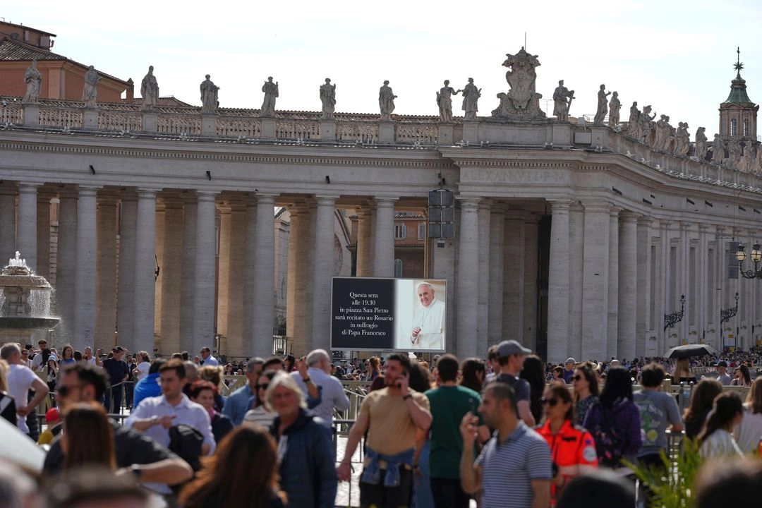 La gente camina por la Plaza de San Pedro del Vaticano mientras una pantalla gigante anuncia que se ha programado un rezo del rosario por el papa Francisco para las 19:30 h del lunes 21 de abril de 2025. Foto