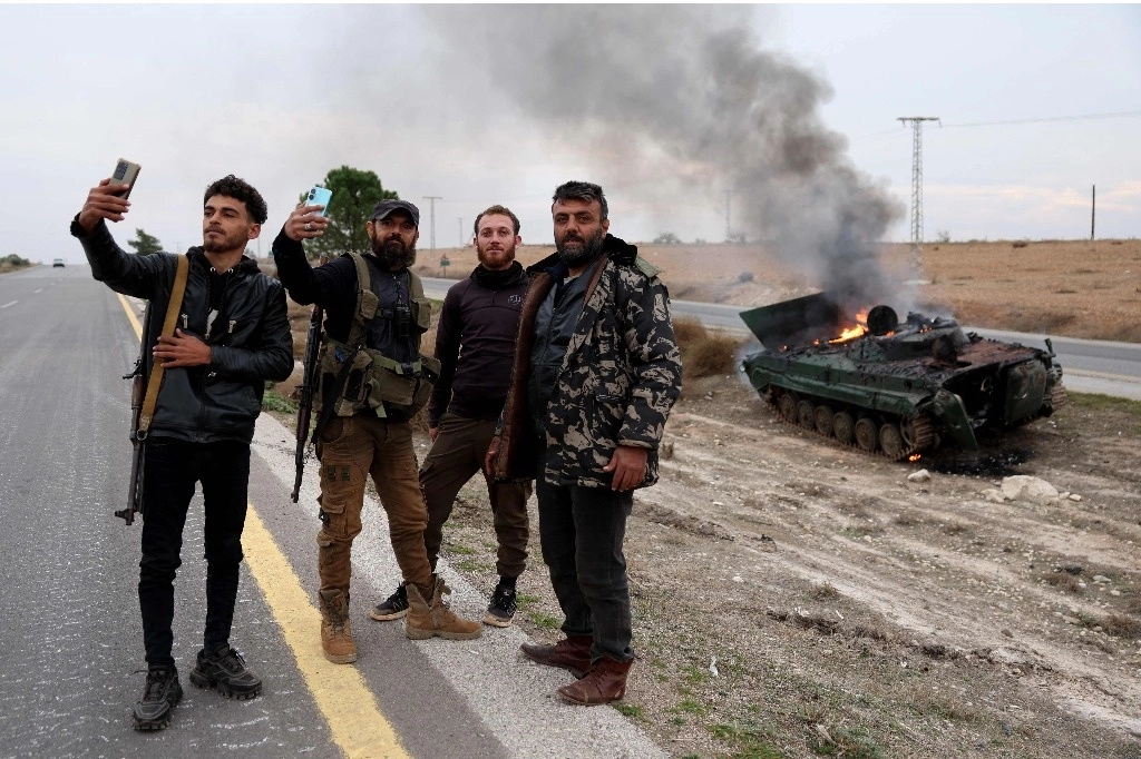 Hombres armados posan para fotografías cerca de un vehículo militar perteneciente a las fuerzas del régimen sirio y confiscado por las fuerzas antigubernamentales, en llamas después de ser alcanzado por las fuerzas del régimen, en la gobernación de Hama, el 7 de diciembre de 2024. Foto Afp