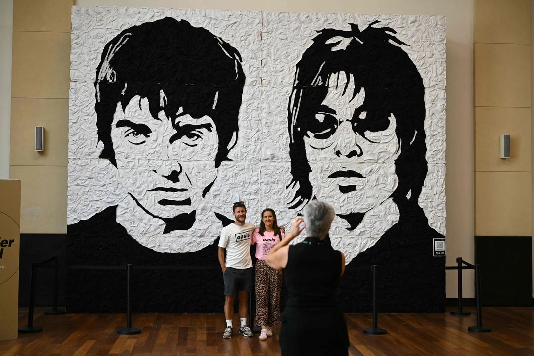 La gente posa para una foto frente a una enorme obra de arte de la banda de rock británica Oasis, llamada "The Wonder Wall", creada por Nathan Wyburn, antes del concierto inaugural de su esperada gira en Cardiff, el 4 de julio de 2025. Foto 