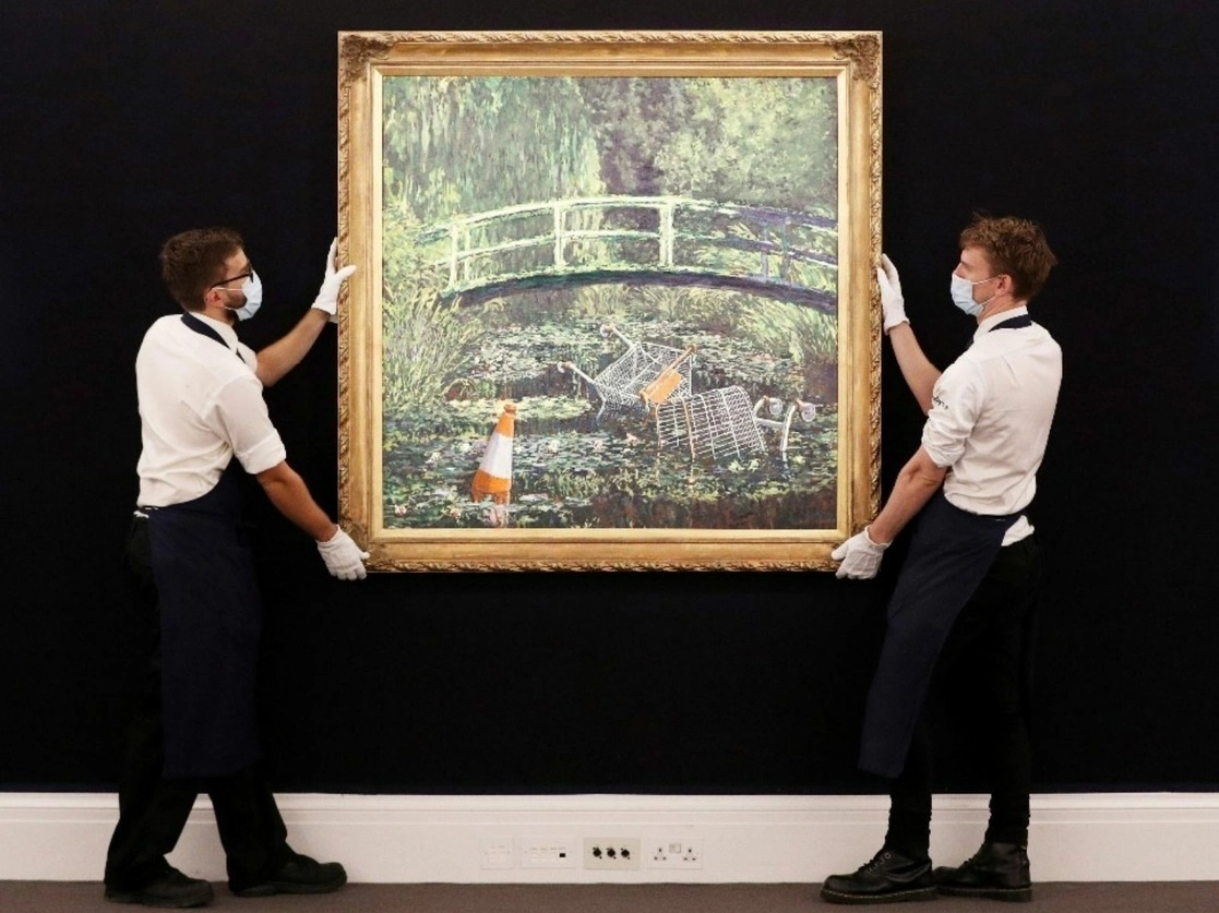 Dos técnicos manejan la obra de Banksy, 'Muéstrame el Monet' en exhibición en Sotheby's en el centro de Londres. La reinterpretación contemporánea de Banksy de la representación de Claude Monet del puente japonés en su jardín en Giverny será parte de una subasta transmitida en vivo por Sotheby's. Foto AP 
